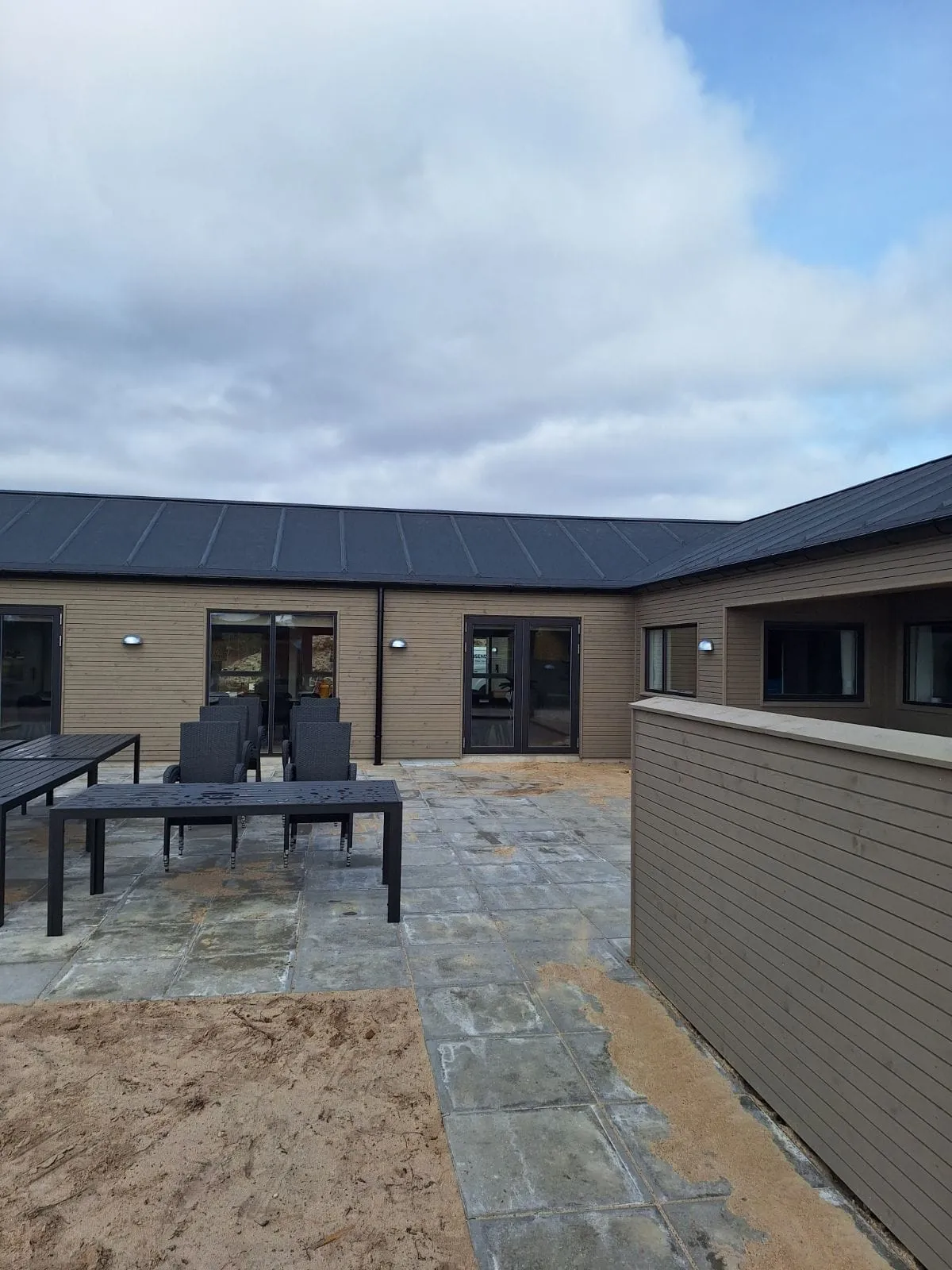 Billedet viser en moderne bolig med træfacade og mørkt tag set fra en rummelig flisebelagt terrasse.