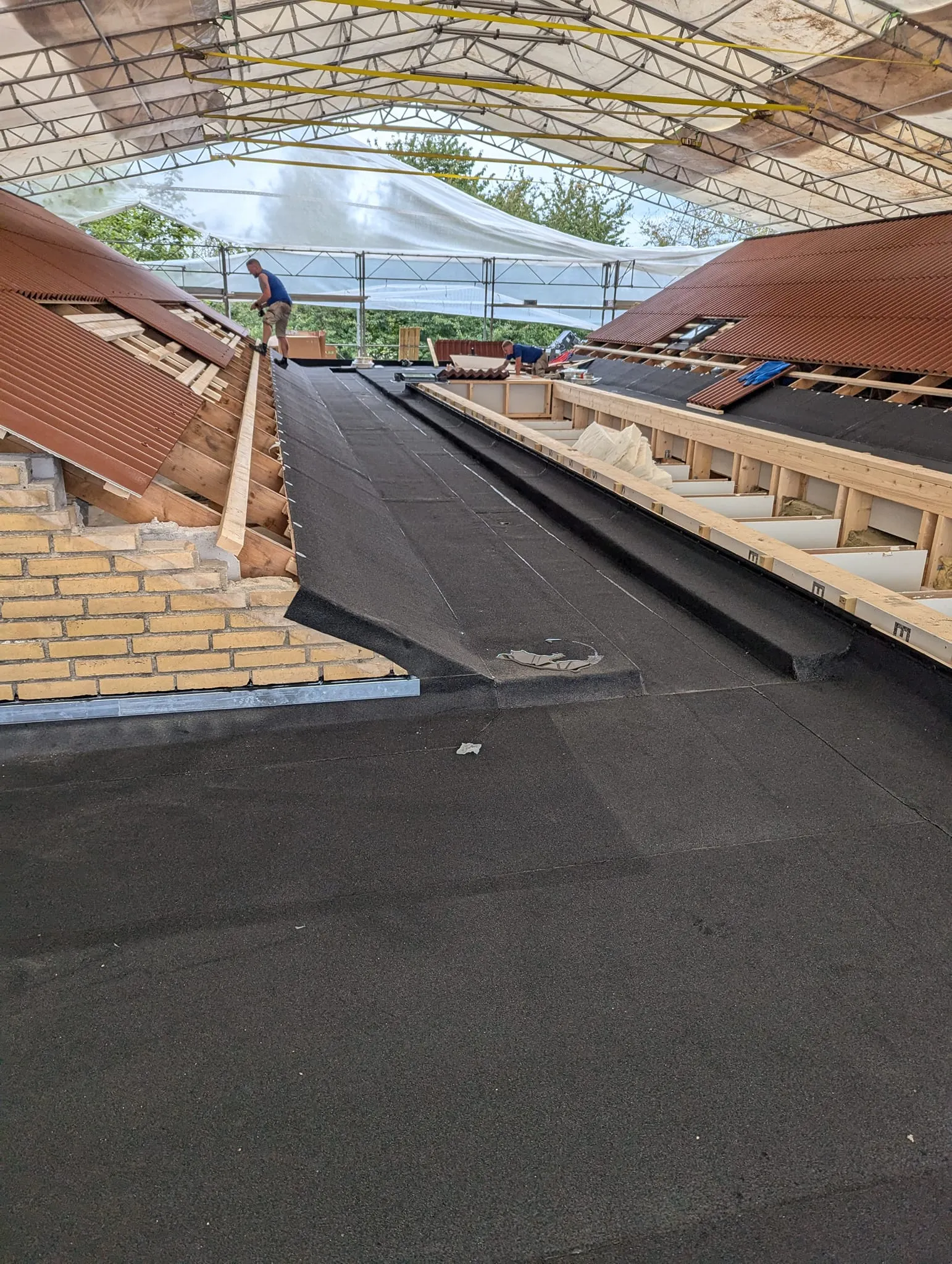 Billedet viser to håndværkere, der arbejder på en tagrenovering under en stor, midlertidig teltkonstruktion.